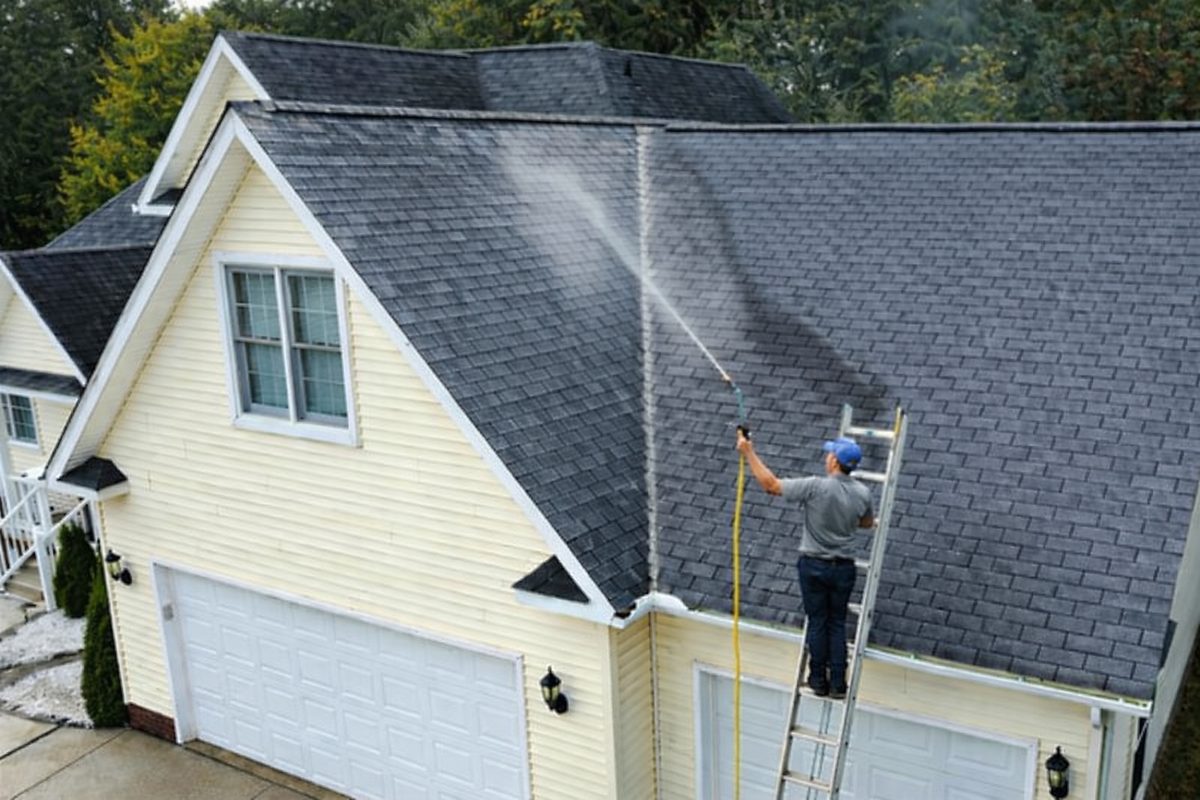 A worker power washing a roof from a ladder, removing dirt and debris.